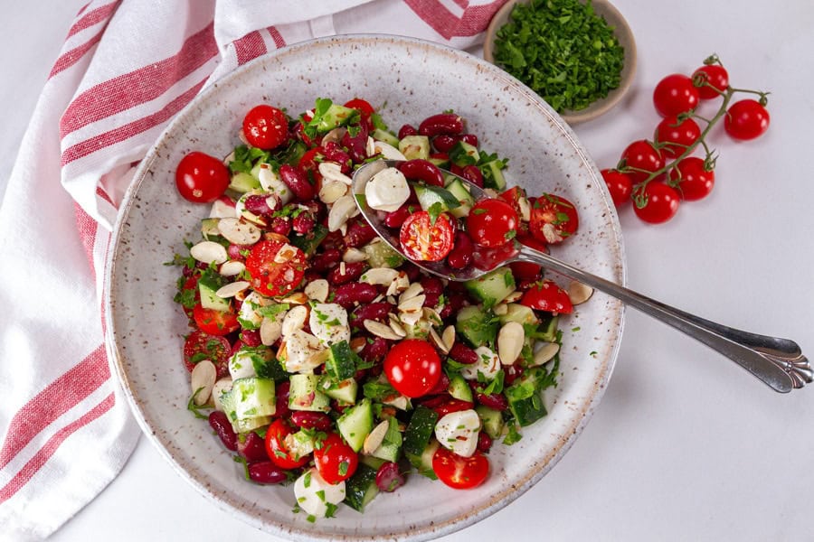 A bowl of salad with cherry tomatoes, kidney beans, cucumber, mozzarella balls, sliced almonds, and chopped herbs, with a fork and a napkin nearby.