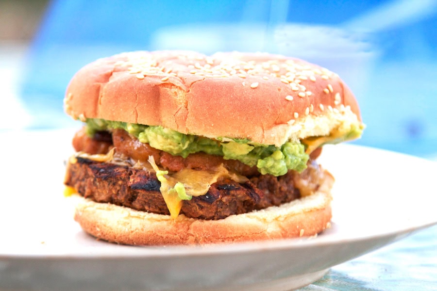 A sesame seed hamburger bun filled with a grilled patty, melted cheese, guacamole, and sauce, served on a white plate.