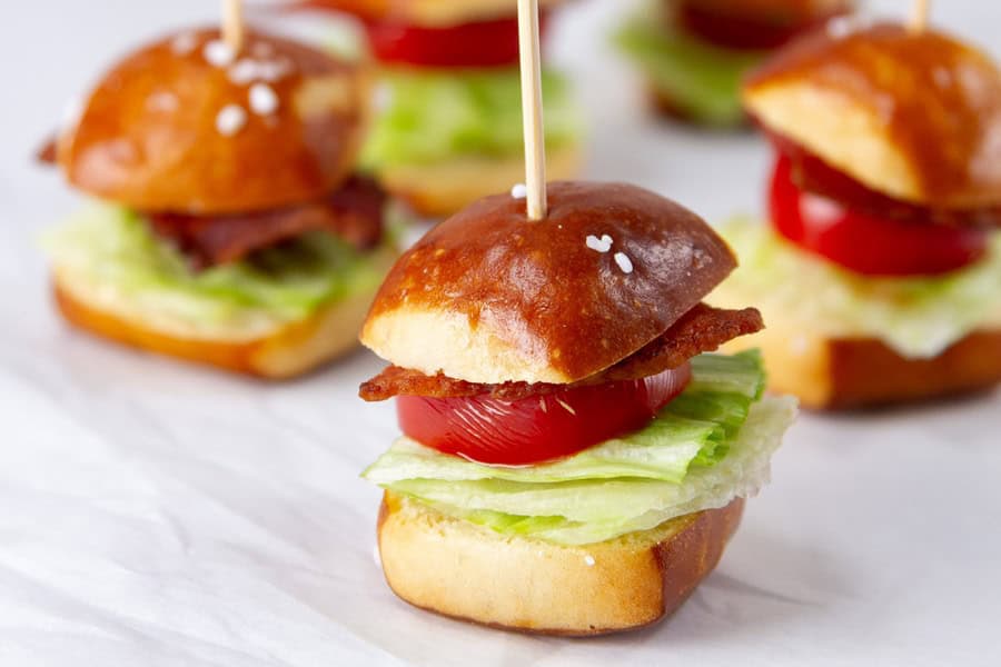 Mini sandwiches with lettuce, tomato, and bacon on small pretzel buns, each held together with a toothpick, arranged on a white surface.