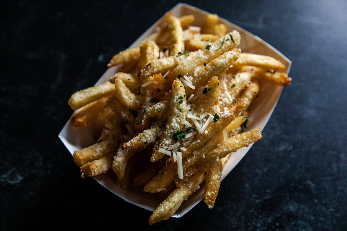 A serving of French fries topped with grated cheese and herbs in a white paper tray on a dark surface.