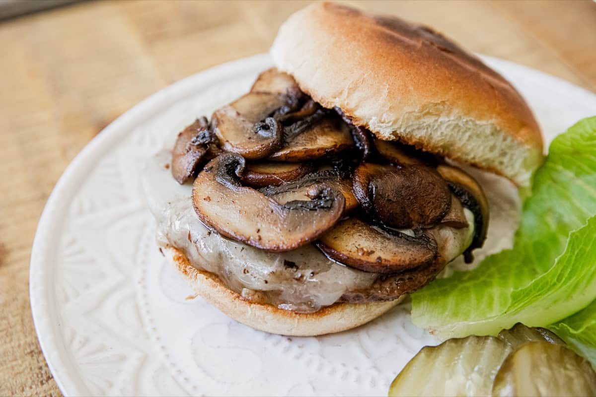 A cheeseburger topped with sautéed mushrooms on a bun, served on a white plate with lettuce and pickle slices on the side.