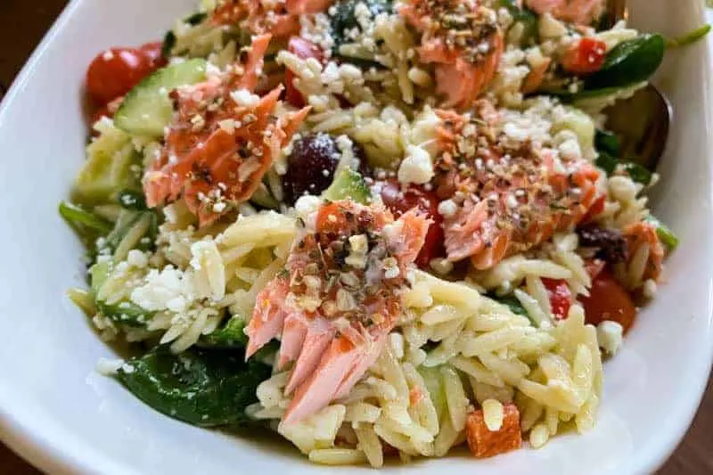 A close-up of a bowl of orzo pasta salad with spinach, cherry tomatoes, olives, feta cheese, and pieces of salmon topped with herbs.