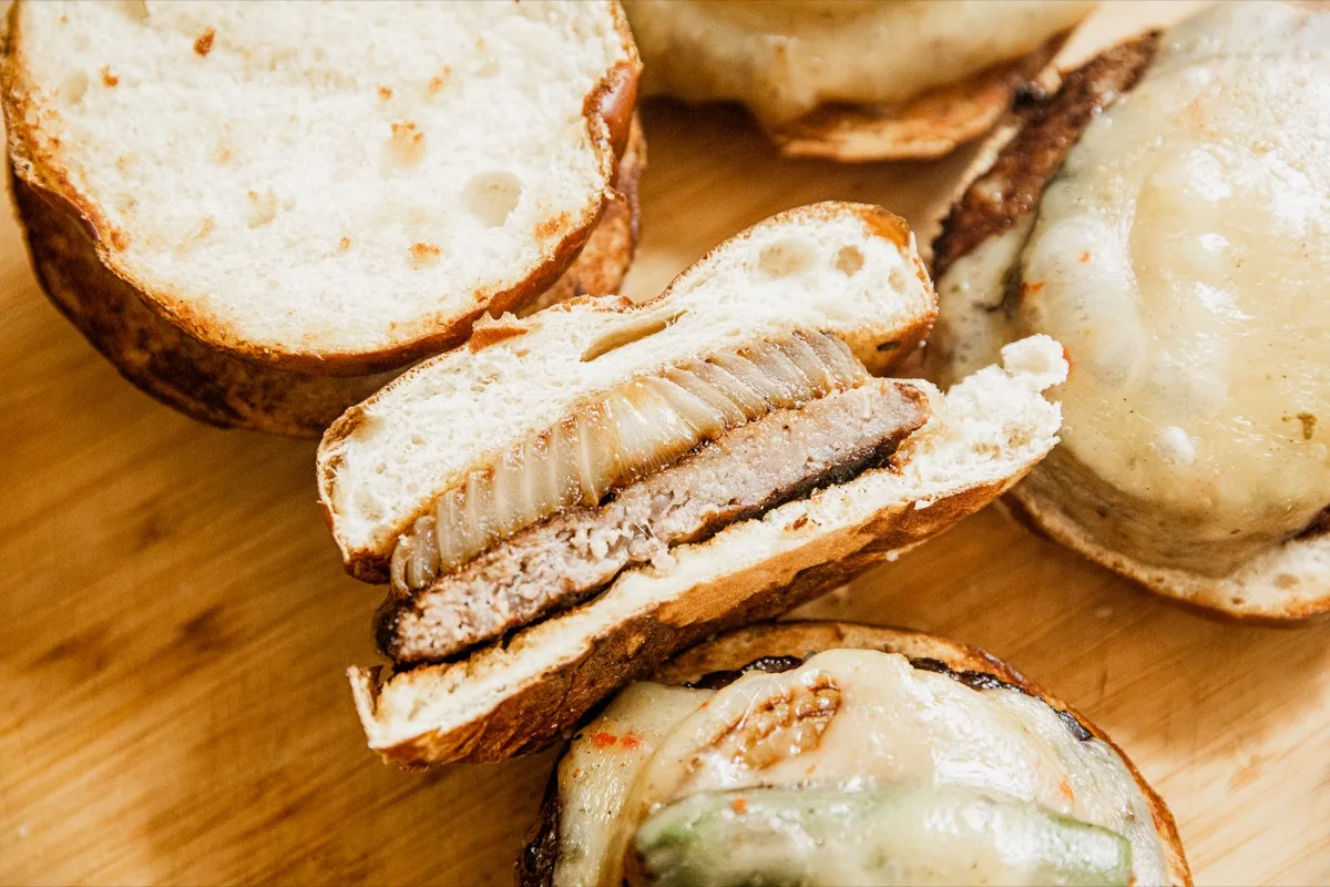 A sliced burger on a wooden surface reveals a beef patty topped with cooked onions, placed inside a bun. Other burgers with melted cheese are visible nearby.
