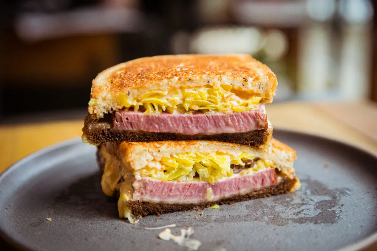 A close-up of a sandwich cut in half, featuring thick slices of meat, shredded lettuce or sauerkraut, and melted cheese on toasted bread, served on a gray plate.