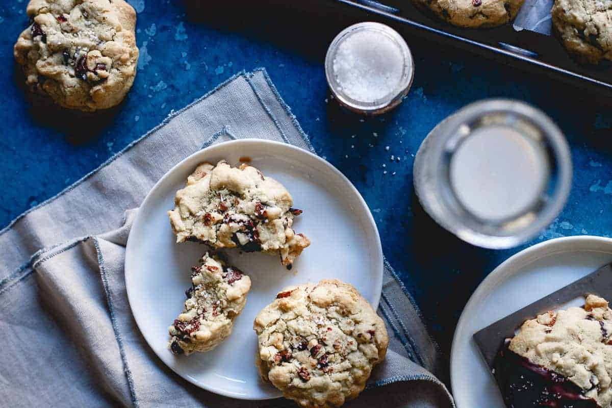 A plate with chocolate chip cookies, one broken in half, sits on a napkin. Nearby are a glass of water, a jar of salt, and a baking tray with more cookies on a blue surface.