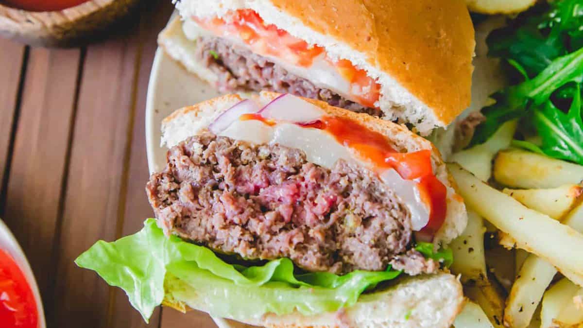 A close-up of a hamburger cut in half, showing lettuce, onion, tomato, ketchup, and a beef patty on a plate with French fries and leafy greens.