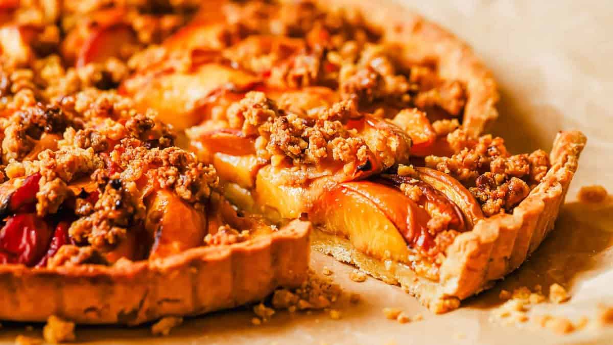 A close-up of a peach crumble tart with a slice being removed, showing a golden crust, baked peaches, and crumbly topping.