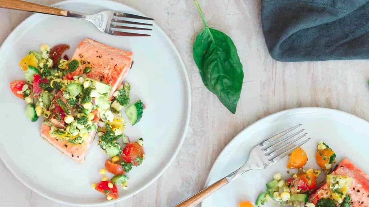 Two plates with grilled salmon fillets topped with a colorful vegetable salsa, each with a fork; a fresh basil leaf and a dark napkin are nearby.