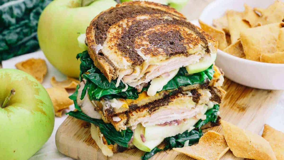 A stacked sandwich with leafy greens, sliced turkey, cheese, and apple slices on swirled bread, served on a wooden board with apple and chips on the side.