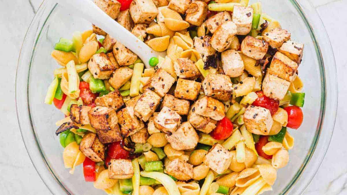 A glass bowl filled with pasta shells, chopped grilled tofu, cherry tomatoes, green bell pepper, and peas, with a white serving spoon.