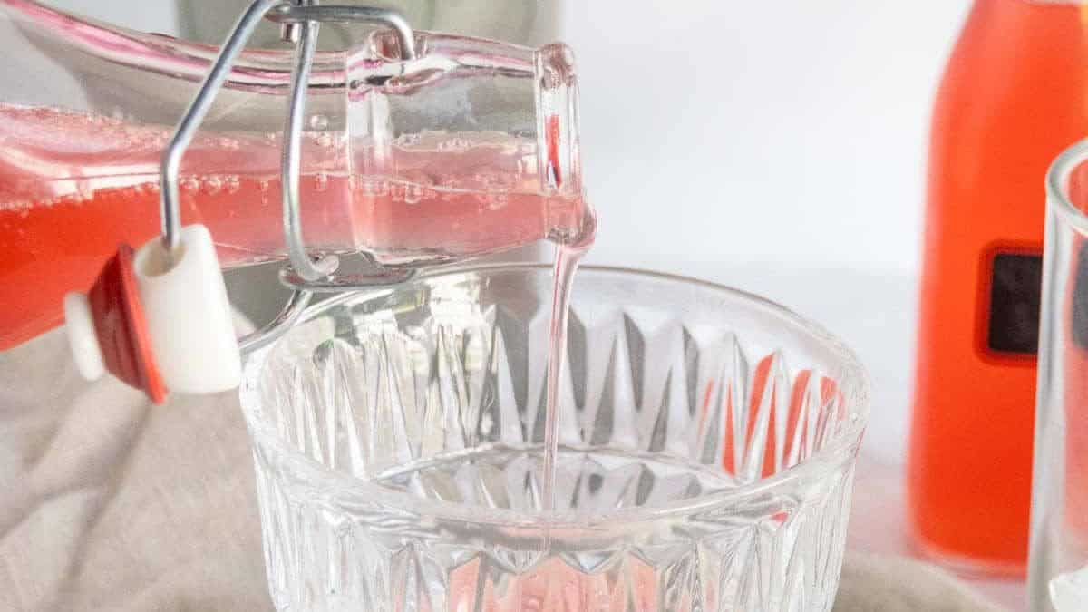 A hand pours pink liquid from a glass bottle into a stemmed crystal glass, with bottles and a beige cloth in the background.