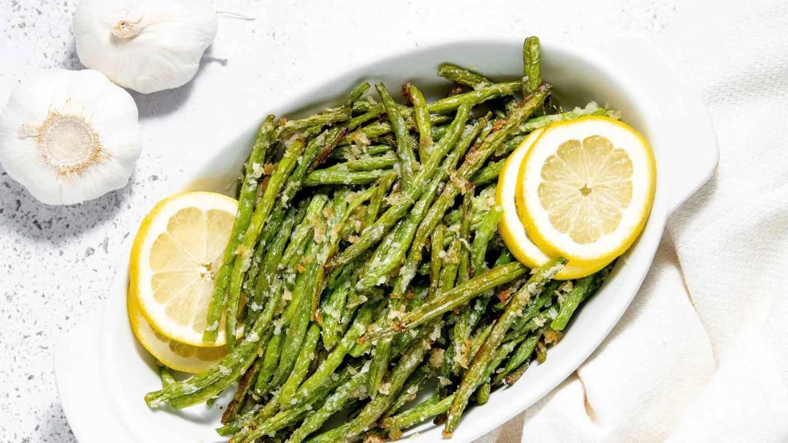 A white dish filled with roasted green beans garnished with grated cheese and two lemon slices, next to two garlic bulbs on a white surface.