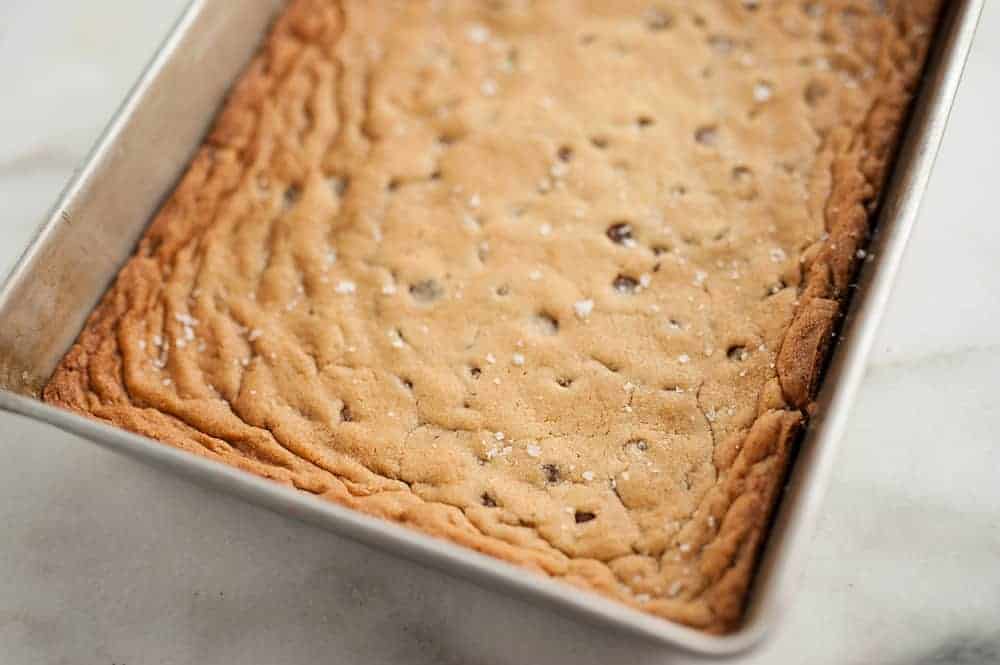 A rectangular pan of baked cookie bars with a golden-brown top and sprinkled sea salt, sitting on a white surface.