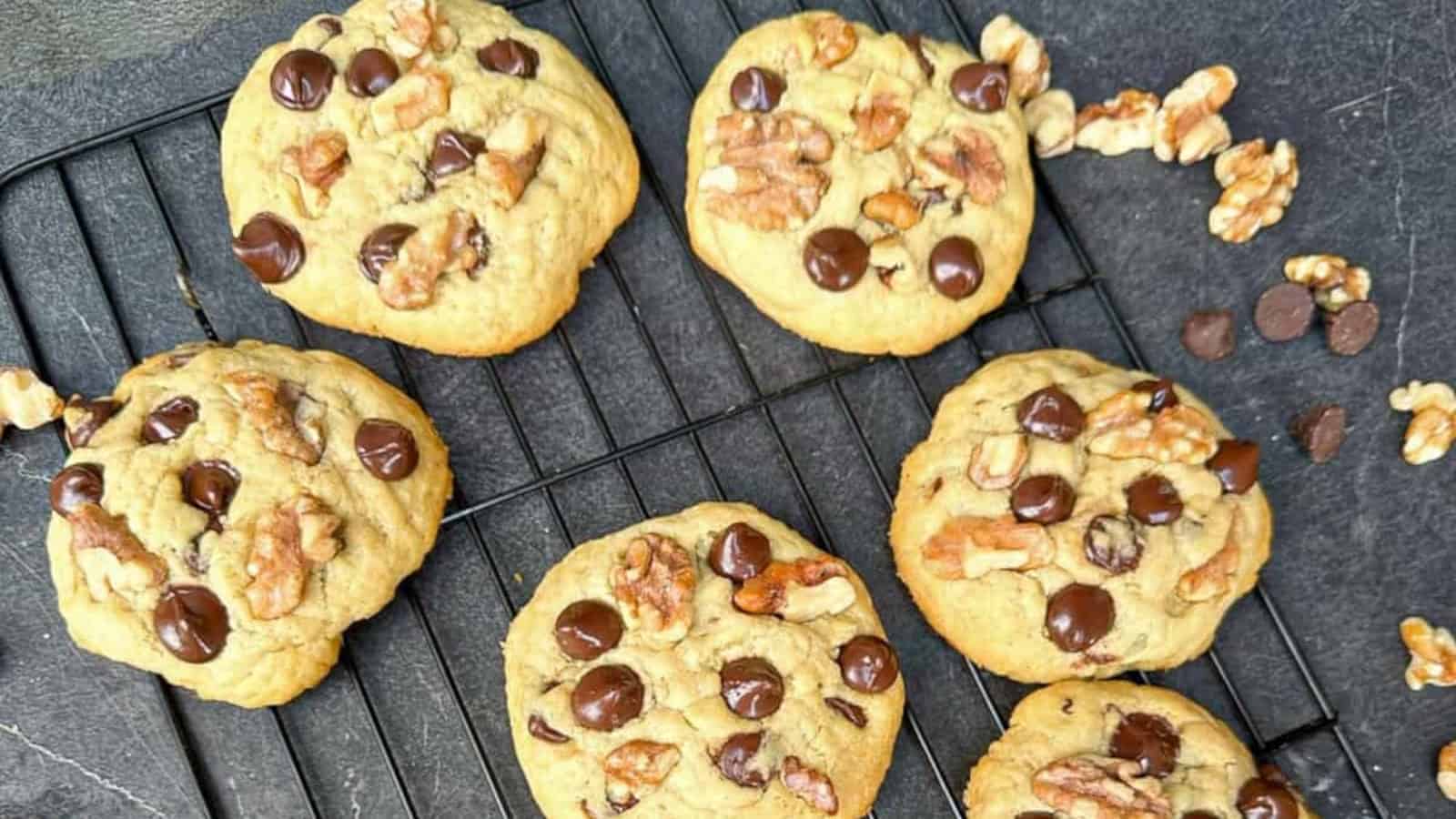 Six chocolate chip and walnut cookies are cooling on a black wire rack, with extra walnut pieces and chocolate chips scattered nearby on a dark surface.