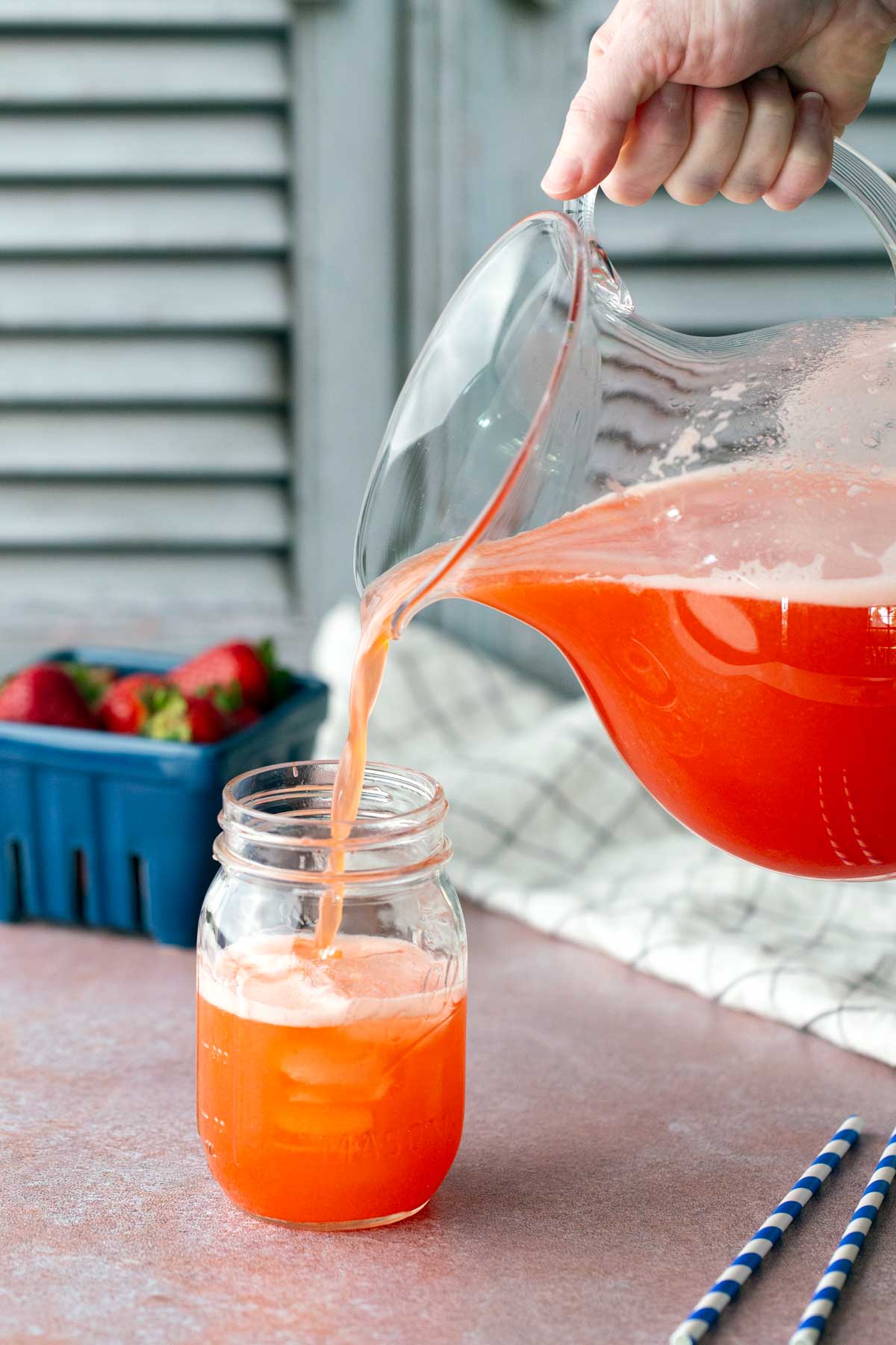 A hand pours bright orange-red juice from a glass pitcher into a mason jar; strawberries and striped straws are nearby.