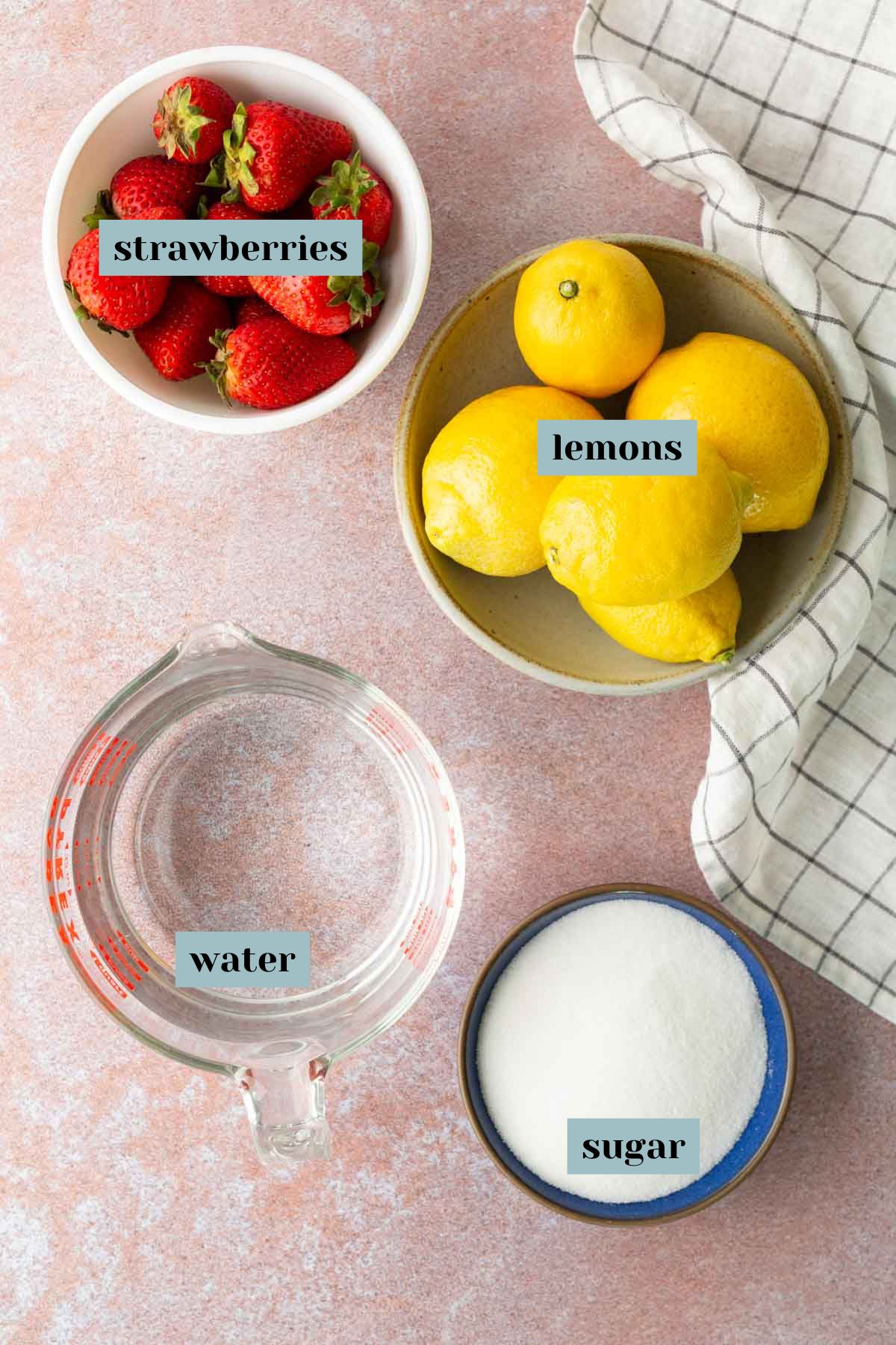 Four labeled bowls on a countertop containing strawberries, lemons, water, and sugar, arranged next to a checkered cloth.
