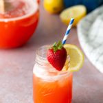 A mason jar of strawberry lemonade with ice, garnished with a lemon slice and whole strawberry, and a blue-striped straw; pitcher and fruit in the background.
