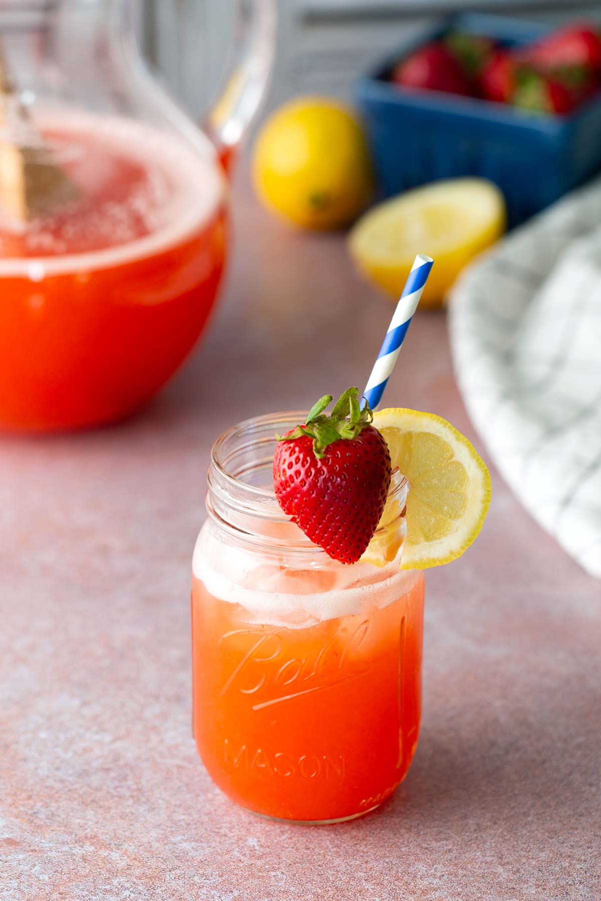 A mason jar of strawberry lemonade with ice, garnished with a lemon slice and whole strawberry, and a blue-striped straw; pitcher and fruit in the background.