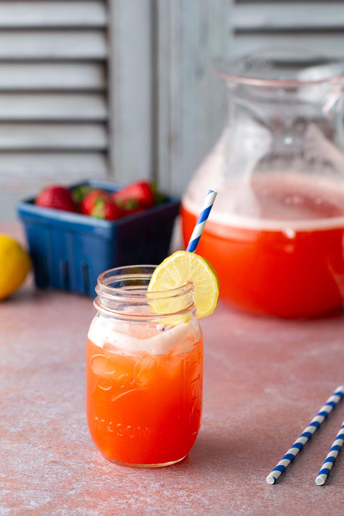 A mason jar filled with a red drink, garnished with a lemon slice and a striped straw. A pitcher of the same drink, strawberries, and lemons are in the background.