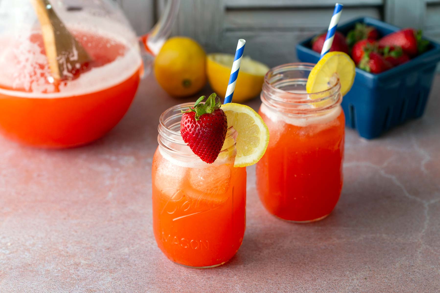 Two mason jars filled with iced strawberry lemonade, garnished with lemon slices and a strawberry, sit on a table with a pitcher and fresh strawberries in the background.