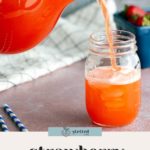 A glass pitcher pours strawberry lemonade into a mason jar on a pink surface, with two striped straws and a container of strawberries in the background.