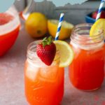 Two mason jars filled with strawberry lemonade, ice, striped straws, and garnished with lemon slices and strawberries. A pitcher and lemons are in the background.