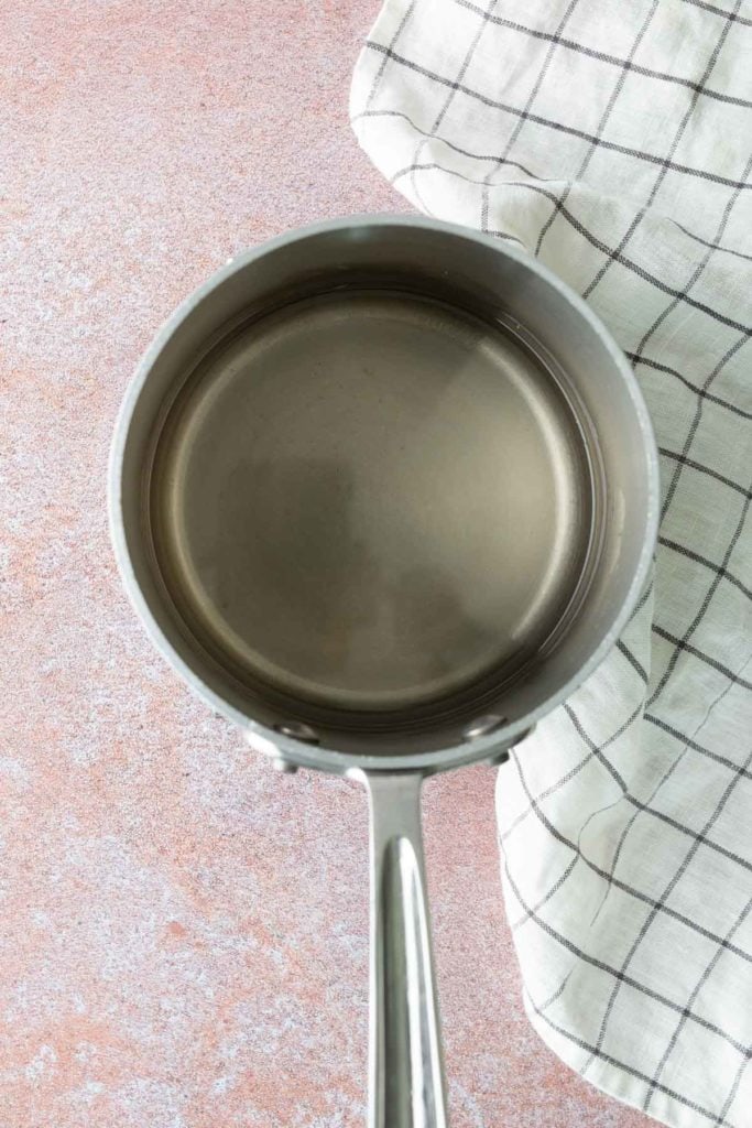 A stainless steel saucepan filled with water sits on a pink countertop next to a folded white and black checkered kitchen towel.