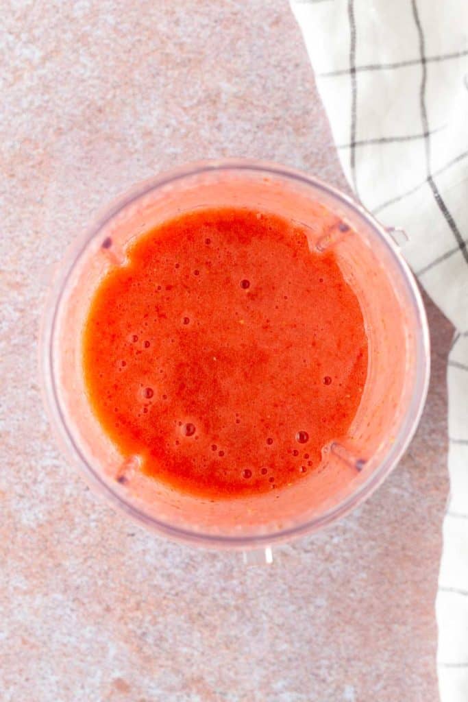 A top-down view of a blender cup filled with a smooth red liquid, likely a fruit smoothie, on a light countertop next to a white cloth with black stripes.