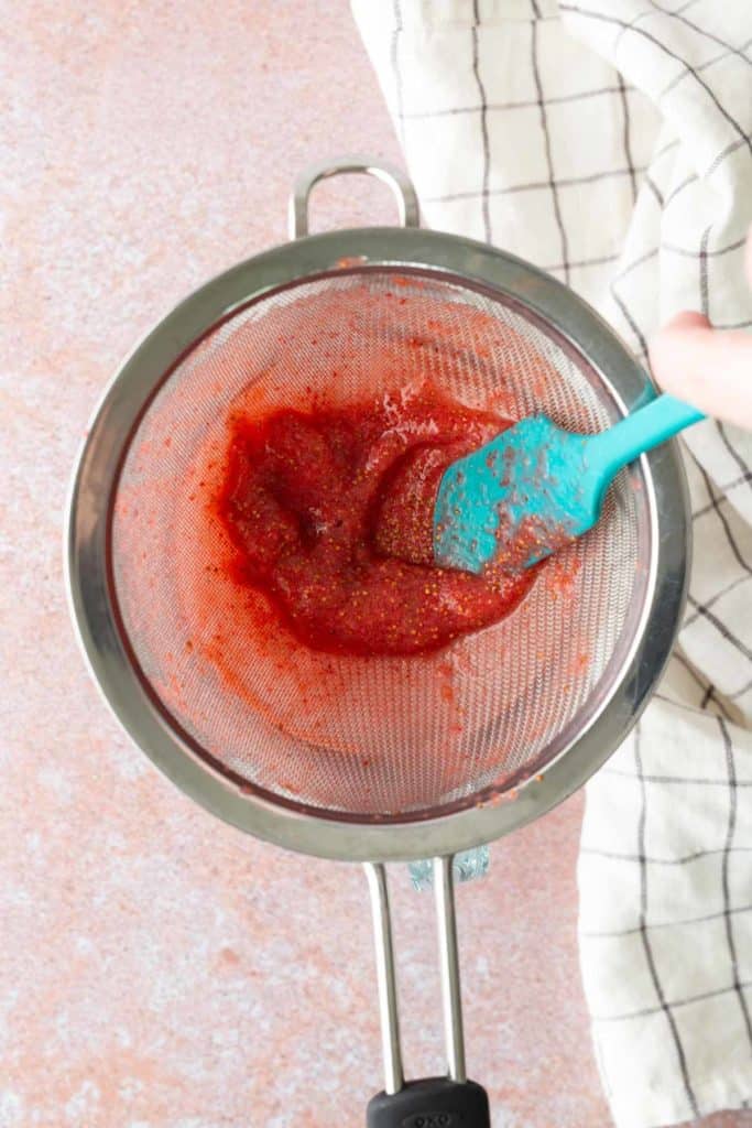 A hand uses a blue spatula to press pureed strawberries through a metal sieve over a bowl, with a white checkered cloth nearby.