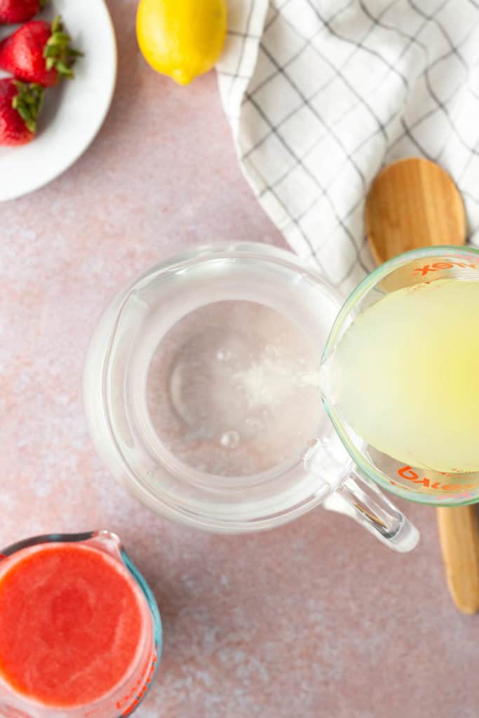 A glass measuring cup pours lemon juice into a clear pitcher. Nearby are a plate of strawberries, a cup of strawberry puree, a lemon, a wooden spoon, and a checkered towel.