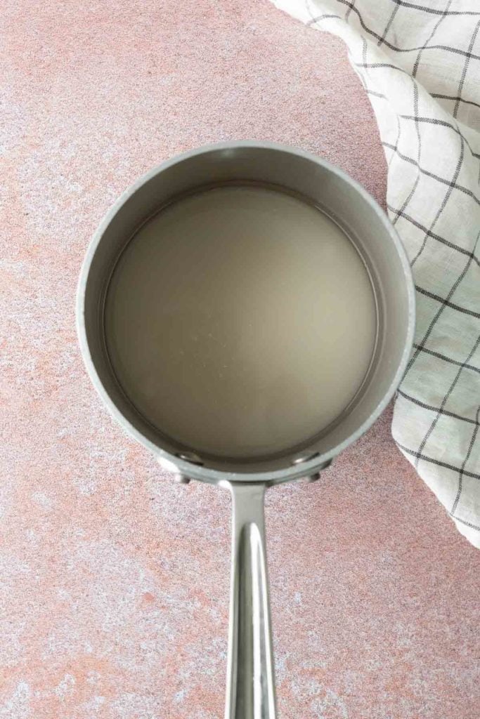 A saucepan filled with a cloudy liquid sits on a pink surface next to a white and black checkered cloth.