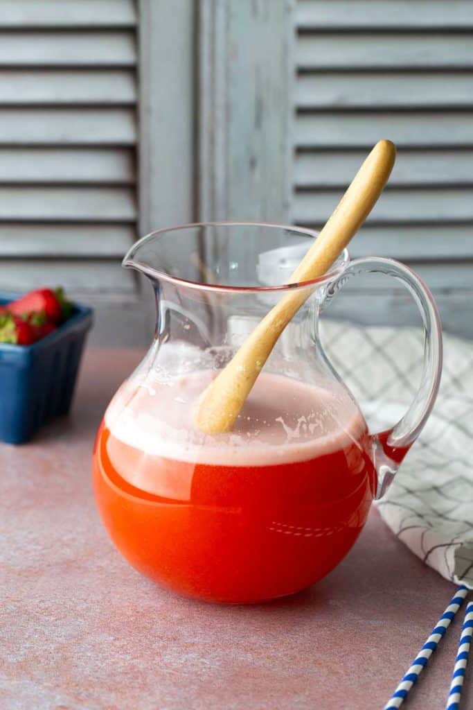 A glass pitcher filled with red strawberry juice and foam, with a wooden spoon inside, sits on a table near a cloth napkin and a basket of strawberries.