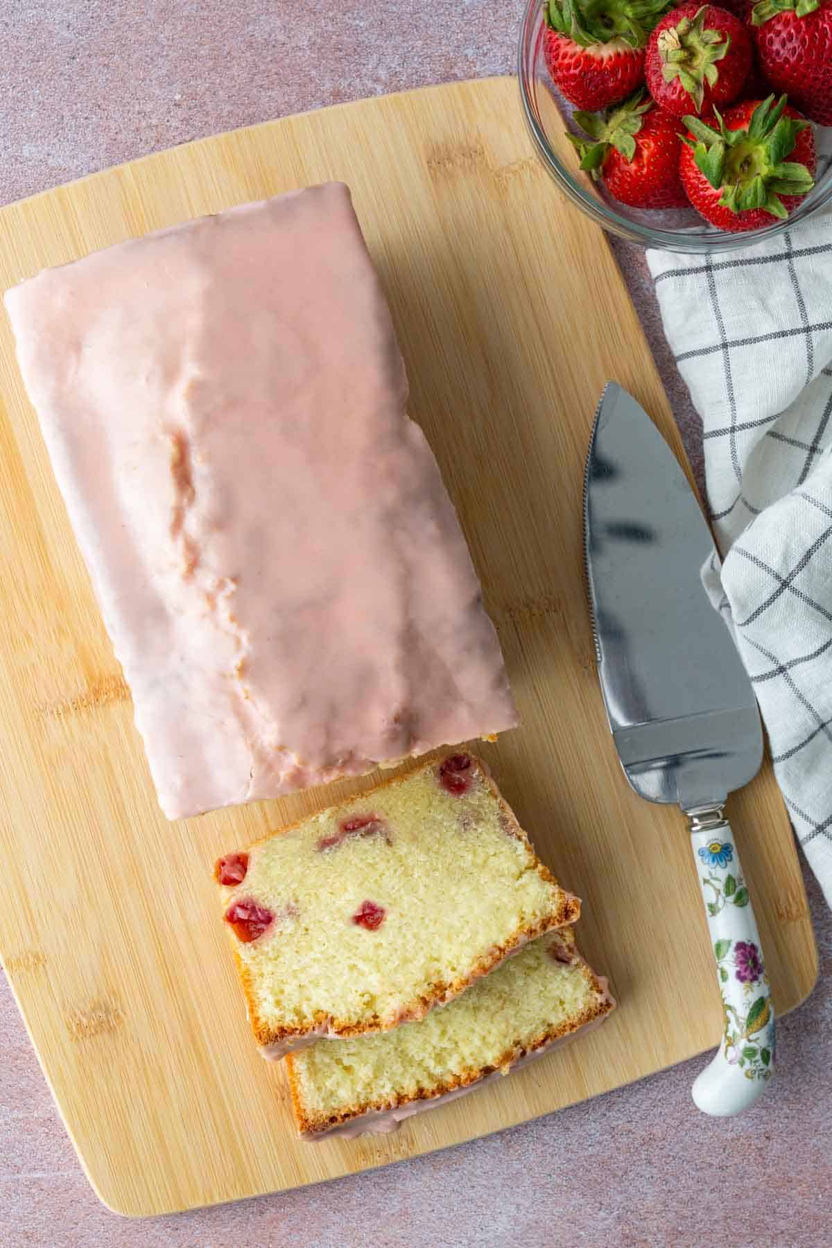 A loaf cake with pink icing sits on a wooden board, two slices cut to show pieces of strawberry inside. A pie server and a bowl of fresh strawberries are nearby.