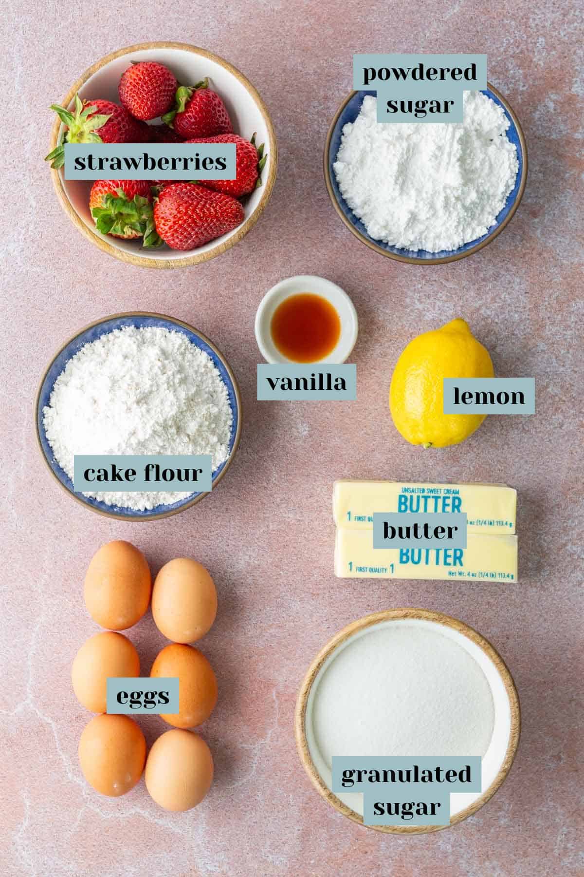 Top-down view of baking ingredients including strawberries, powdered sugar, cake flour, eggs, vanilla extract, lemon, butter, and granulated sugar, each labeled clearly.