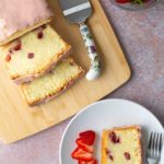 Sliced strawberry loaf cake with pink icing on a wooden board, a serving spatula, fresh strawberries in a bowl, and a plate with cake and sliced strawberries.
