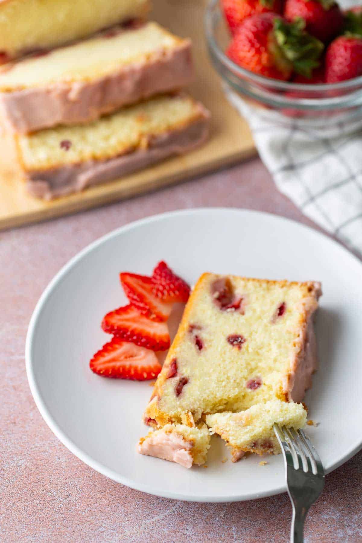 A slice of strawberry loaf cake with icing and a few sliced strawberries on a white plate, with more cake and strawberries in the background.