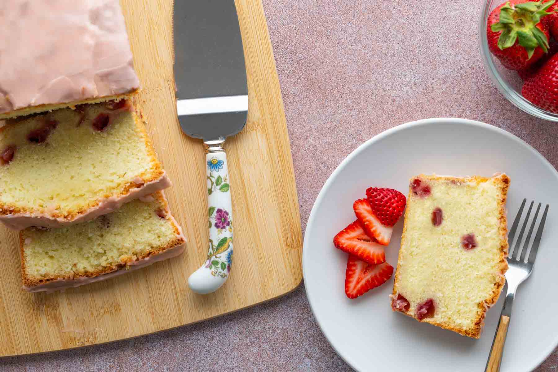 A loaf of strawberry pound cake is sliced on a wooden board; a plate holds a slice with fresh strawberries and a fork, and a bowl of strawberries is nearby.