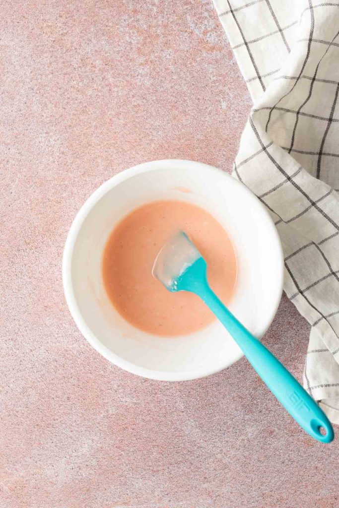 A white bowl with pink sauce and a blue spatula on a light pink surface, next to a white dish towel with black grid lines.