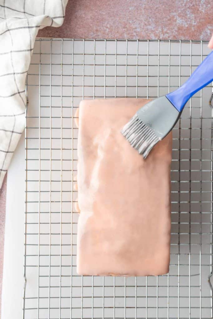 A hand uses a blue pastry brush to apply glaze to a rectangular loaf cake on a wire cooling rack, with a checkered cloth nearby.