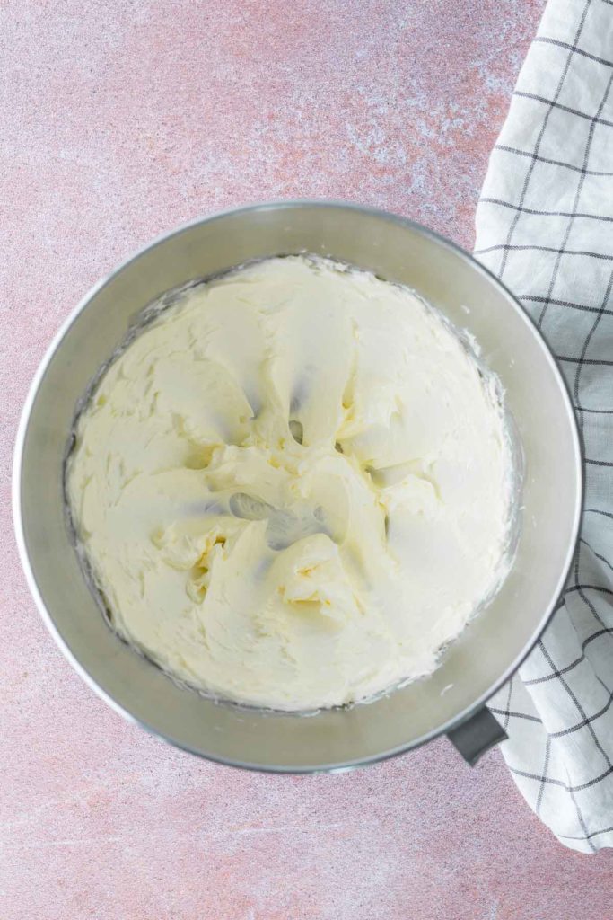 A mixing bowl filled with whipped, creamy buttercream frosting sits next to a white checkered kitchen towel on a pink surface.