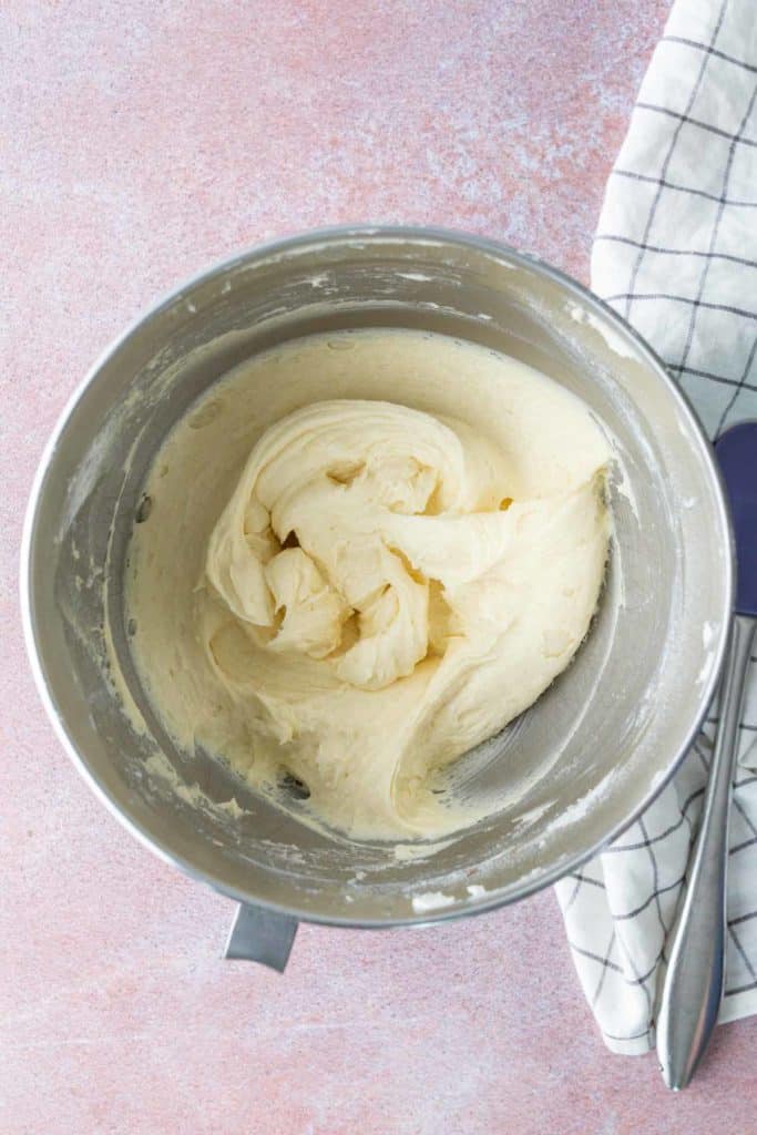 A metal mixing bowl containing thick, creamy dough sits on a light pink surface next to a white kitchen towel with a black grid pattern.
