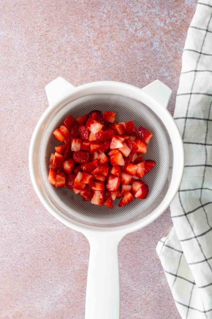 Diced strawberries in a white strainer, placed on a pink surface next to a white and black checkered cloth.