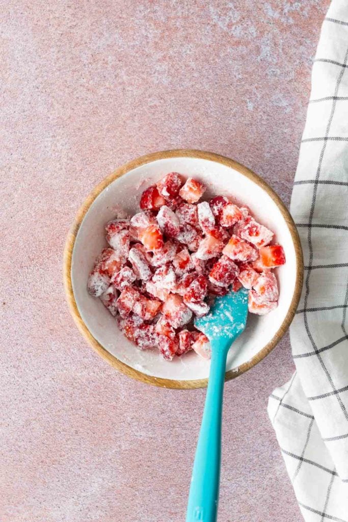 A bowl of chopped strawberries coated in white powder, with a blue spatula, on a pink surface next to a white and black checkered cloth.