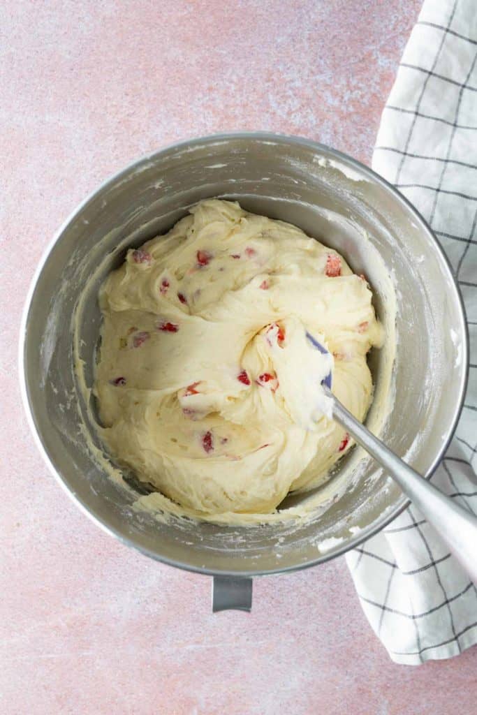A metal mixing bowl filled with creamy batter and red pieces, with a spatula inside. A checkered cloth is partially visible next to the bowl.