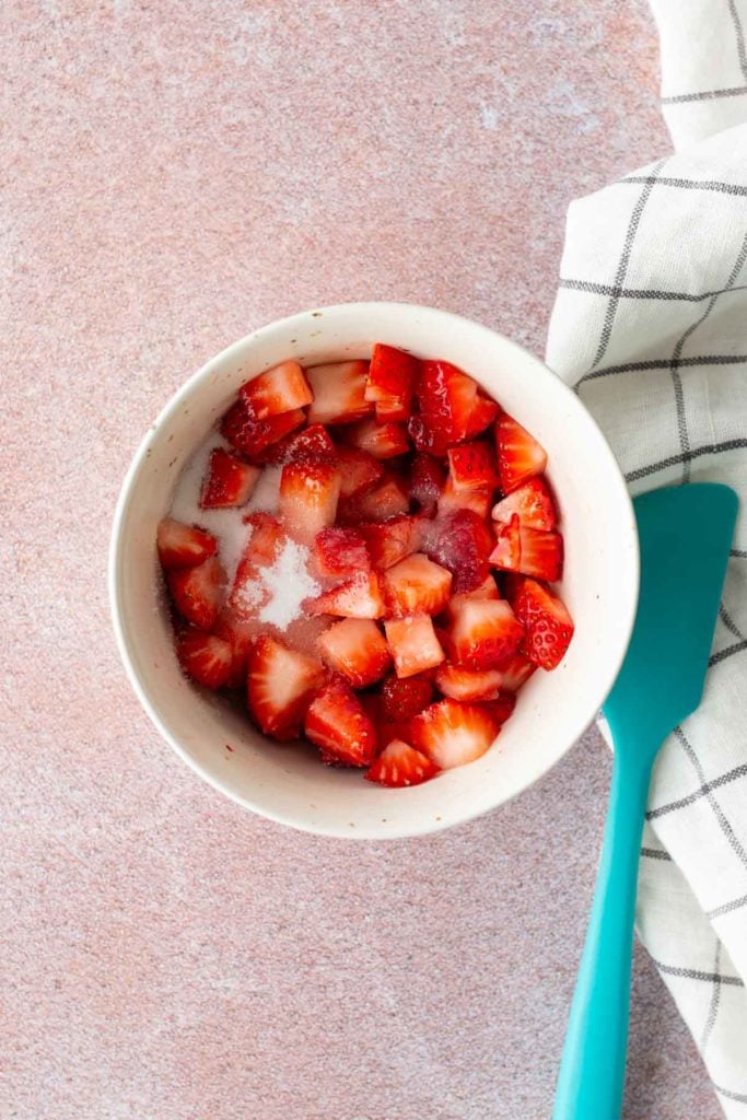 A white bowl filled with chopped strawberries and sugar, next to a blue spatula and a white checkered cloth on a pink surface.