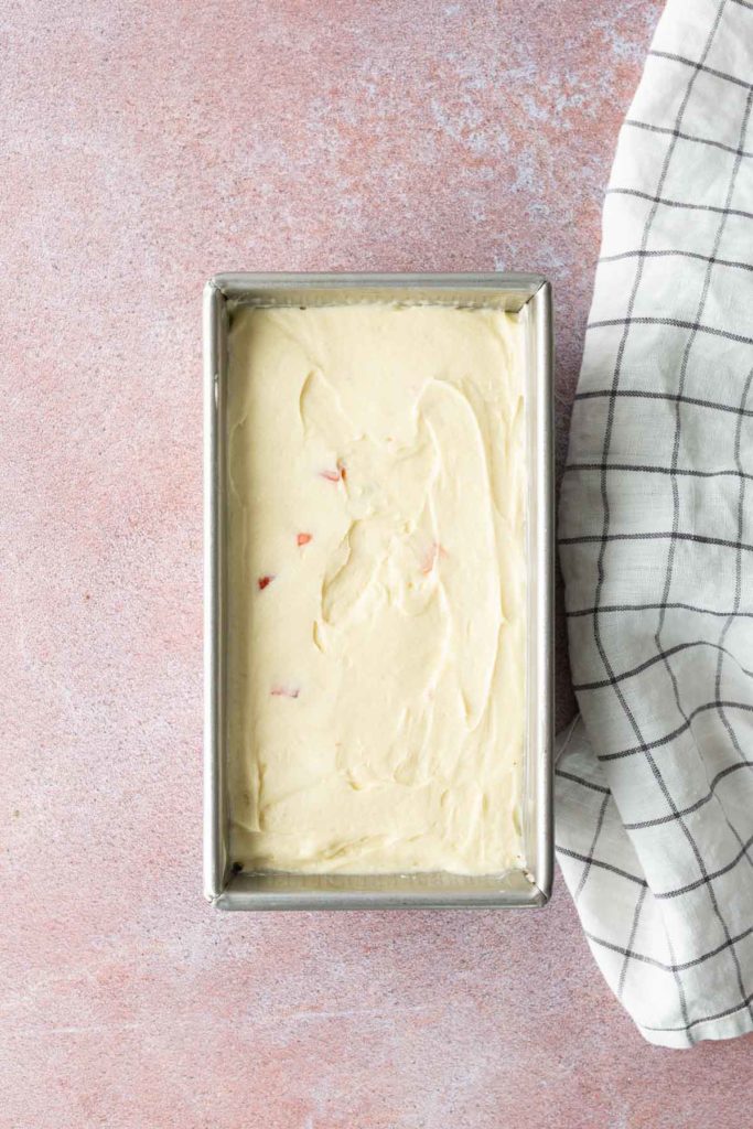 A rectangular metal loaf pan filled with light-colored batter sits on a pink surface next to a white towel with a black grid pattern.