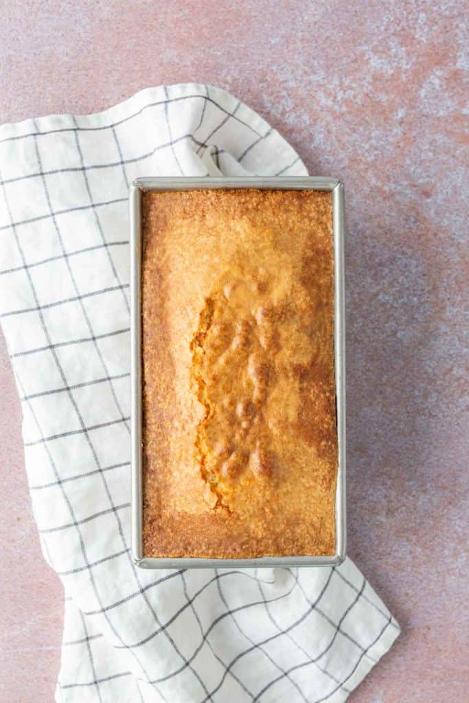 A baked loaf of bread in a rectangular pan sits on a white dish towel with a black grid pattern, on a pinkish countertop.