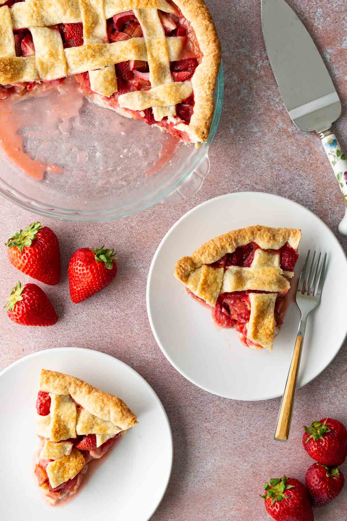 Two slices of strawberry pie with lattice crust on white plates, beside a pie dish with remaining pie, strawberries, a fork, and a pie server on a pink surface.