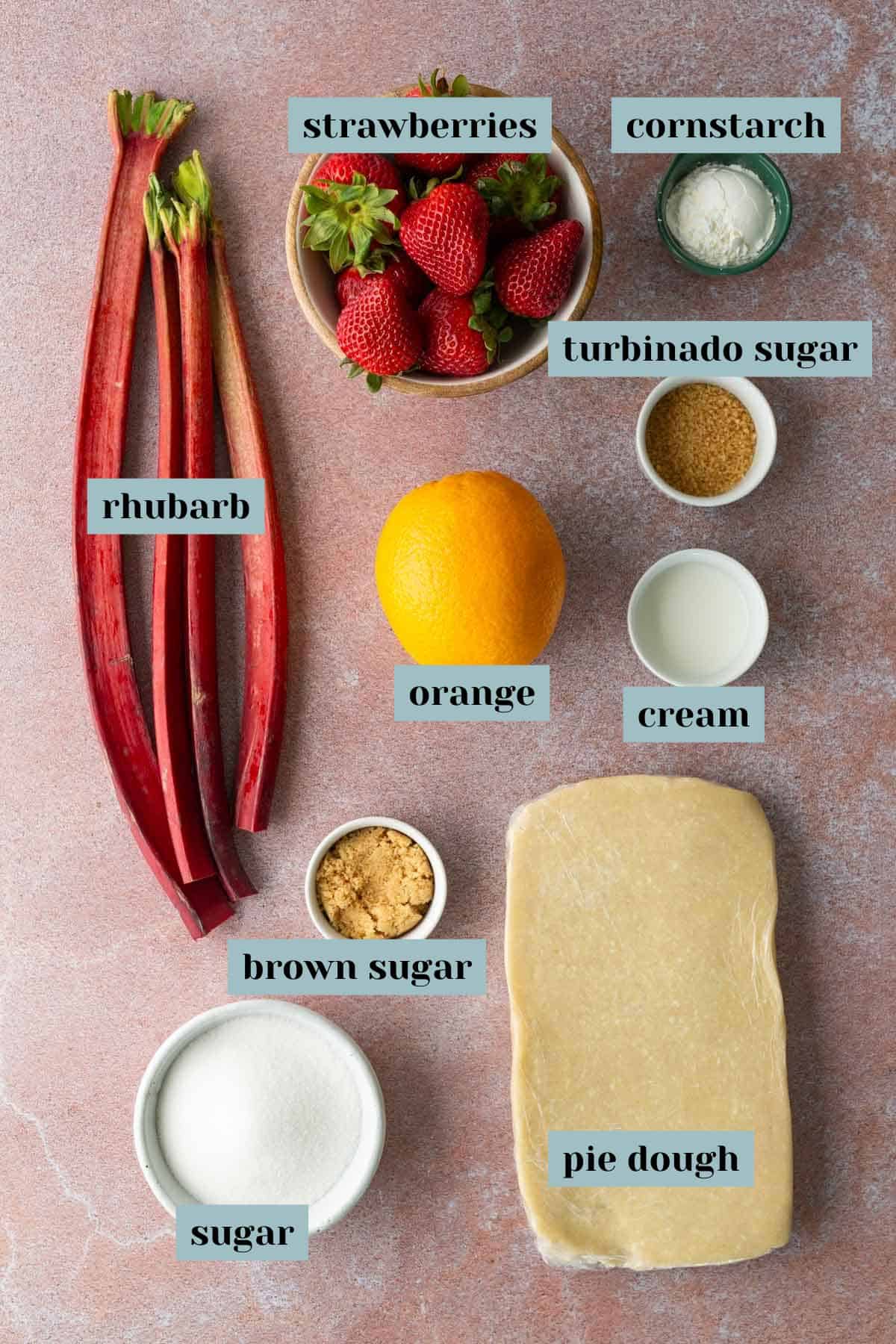 Overhead view of labeled ingredients for a dessert: rhubarb, strawberries, orange, cornstarch, turbinado sugar, cream, brown sugar, pie dough, and white sugar on a pink surface.