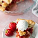 A slice of strawberry pie topped with a scoop of vanilla ice cream, served on a plate with two fresh strawberries and a fork. The rest of the pie is in the background.